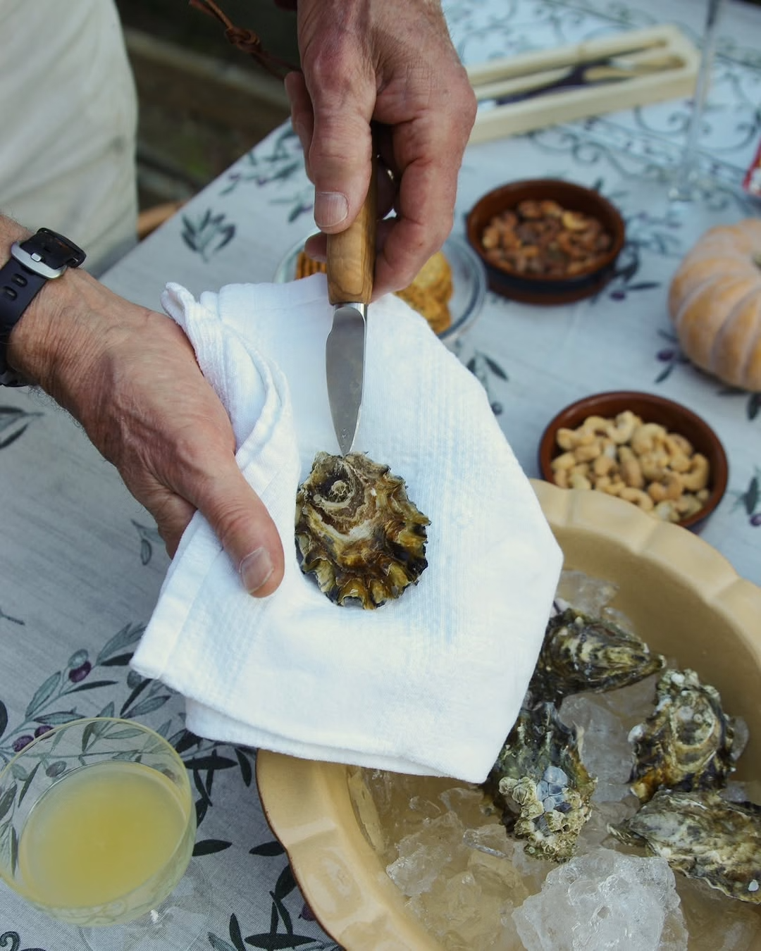 Person opening an oyster with a knife on a white napkin, surrounded by oysters and a bowl of liquid on a floral tablecloth.