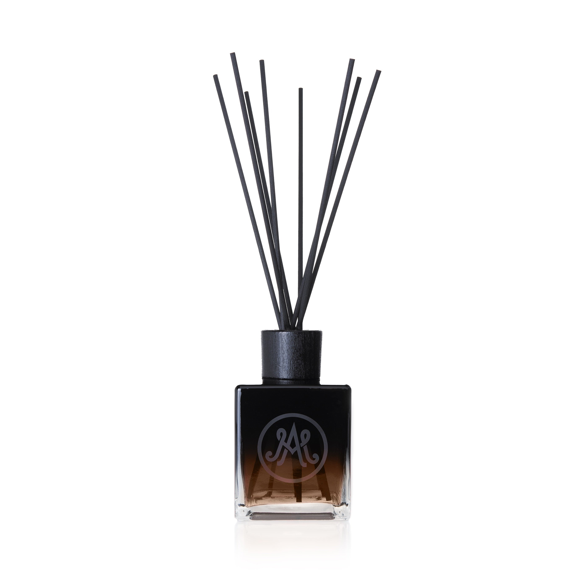 A dark brown diffuser with black reeds is displayed, featuring a sleek black cap and an elegant monogram logo on the front of the bottle. Frankincense, vetiver, leather, tobacco, and ashes make unique, bold fragrance.